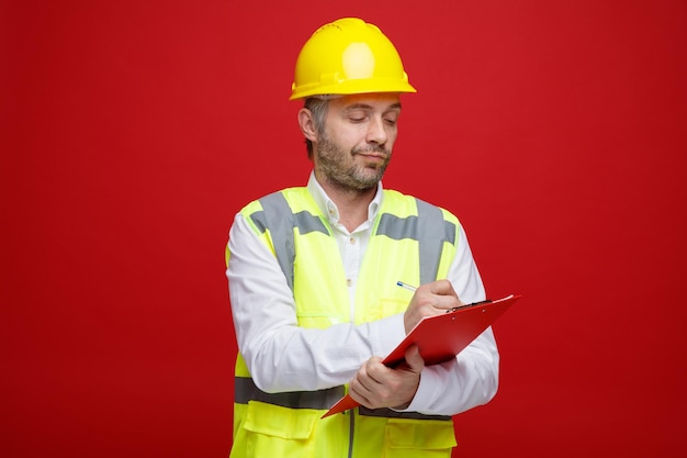 Builder man in construction uniform and safety helmet holding clipboard looking confident making notes standing over red background