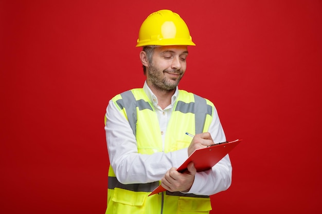 Builder man in construction uniform and safety helmet holding clipboard looking confident making notes standing over red background