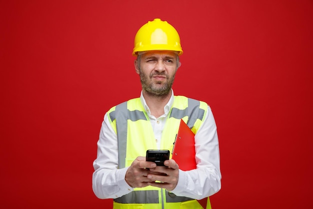 Builder man in construction uniform and safety helmet holding clipboard texting message using smartphone looking displeased making wry mouth standing over red background