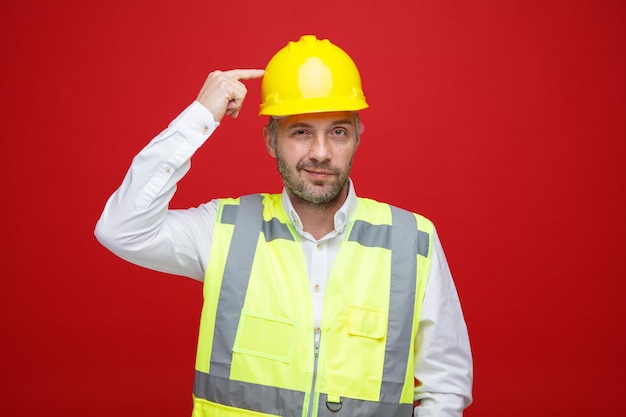 Builder man in construction uniform and safety helmet looking at camera with skeptic smile pointing with index finger at his head standing over red background