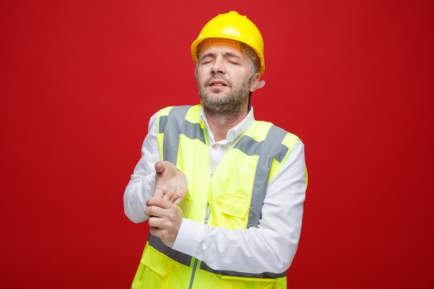 Builder man in construction uniform in safety helmet looking tired and overworked stretching his hands standing over red background