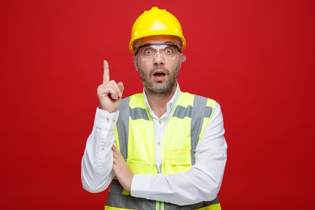 Builder man in construction uniform and safety helmet wearing safety glasses looking at camera being surprised and amazed showing index finger standing over pink background