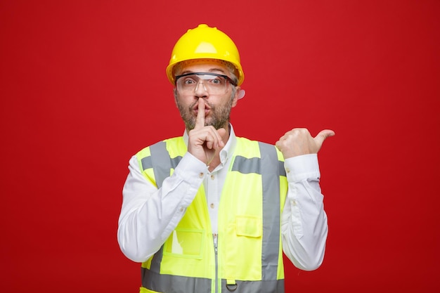 Builder man in construction uniform and safety helmet wearing safety glasses looking at camera making silence gesture with finger on lips pointing with thumb to the side standing over red background