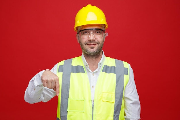 Builder man in construction uniform and safety helmet wearing safety glasses looking at camera smiling pointing with index finger down standing over pink background