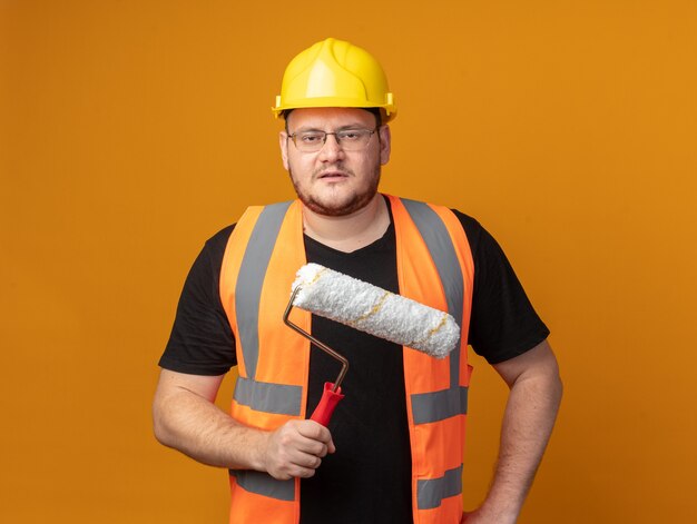 Free photo builder man in construction vest and safety helmet holding paint roller looking at camera with serious face