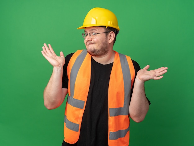 Builder man in construction vest and safety helmet looking at camera confused spreading arms to the sides having no answer standing over green background