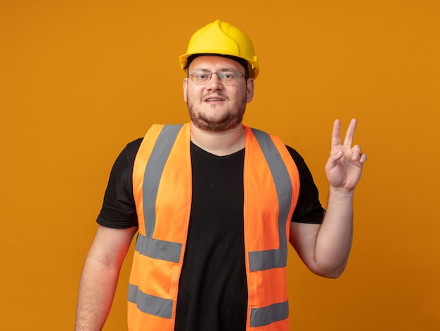 Free photo builder man in construction vest and safety helmet looking at camera smiling confident showing v-sign standing over orange background