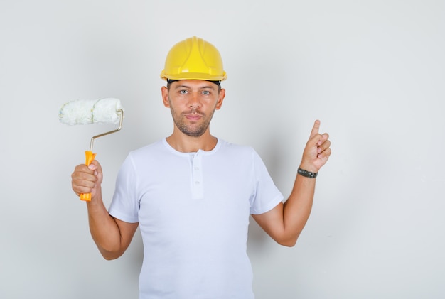 Builder man showing wall and holding paint roller in white t-shirt, helmet, front view