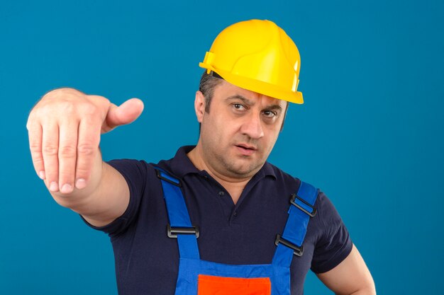 Builder man wearing construction uniform and safety helmet making come here gesture with hand with serious face over isolated blue wall