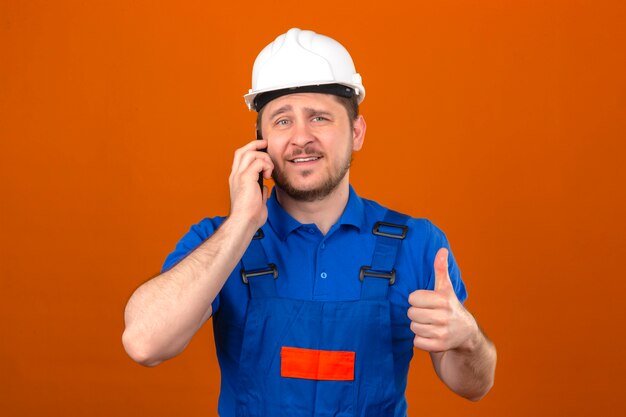 Builder man wearing construction uniform and security helmet talking on mobile phone smiling showing thumb up to camera standing over isolated orange wall