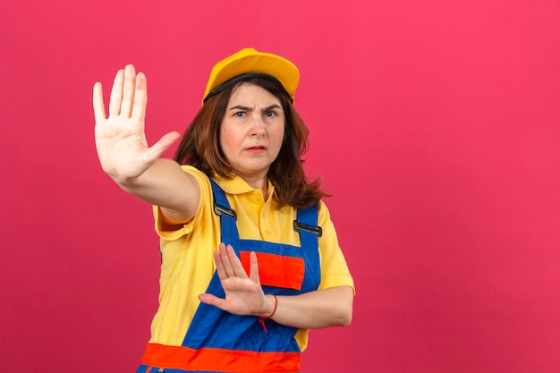 Builder woman wearing construction uniform and yellow cap standing with open hands doing stop sign with serious and confident expression defense gesture over isolated pink wall