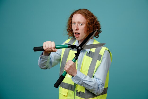 Builder young man in construction uniform holding scissors looking at camera being excited and happy standing over blue background