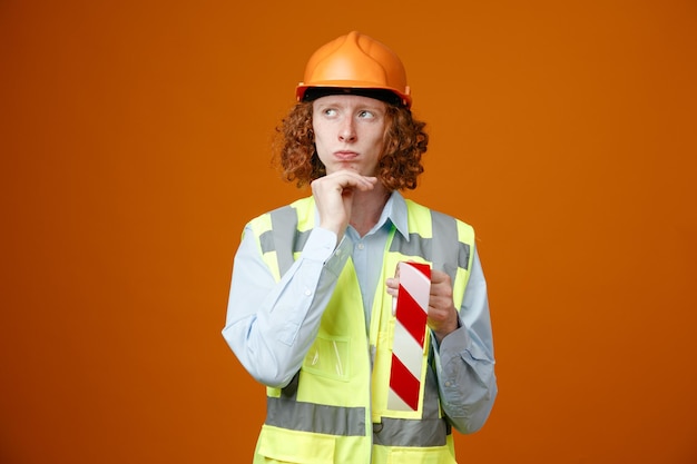 Builder young man in construction uniform and safety helmet holding adhesive tape looking aside with pensive expression thinking with hand on his chin standing over orange background