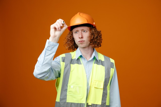 Builder young man in construction uniform and safety helmet holding marker writing something in air with it looking confident standing over orange background