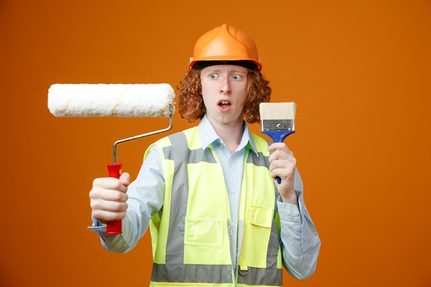 Builder young man in construction uniform and safety helmet holding paint roller and brush looking confused having doubts standing over orange background