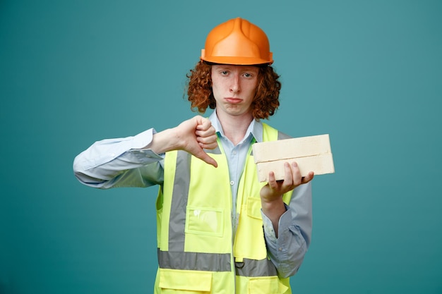 Free photo builder young man in construction uniform and safety helmet holding two bricks looking at camera dissatisfied showing thumb down standing over blue background