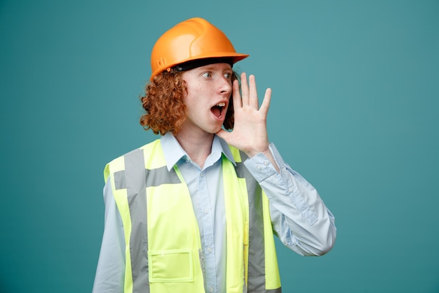 Free Photo builder young man in construction uniform and safety helmet looking aside calling someone with hand near his mouth standing over blue background