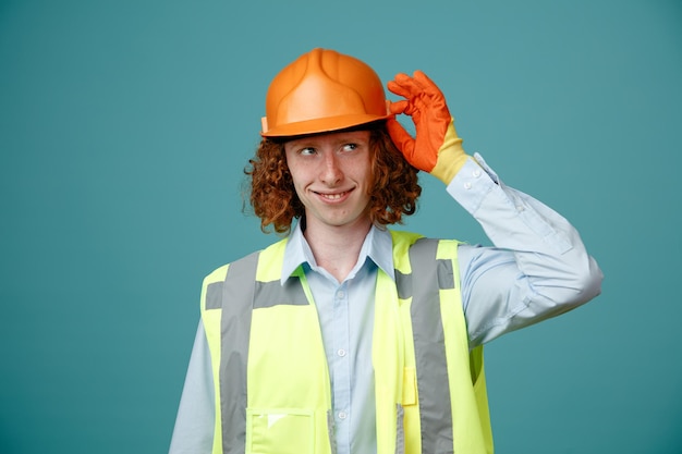 Free photo builder young man in construction uniform and safety helmet wearing rubber gloves looking aside smiling cheerfully happy and positive standing over blue background