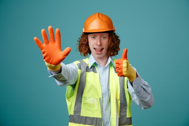 Builder young man in construction uniform and safety helmet wearing rubber gloves looking at camera showing number five with open palm showing thumb up smiling standing over blue background