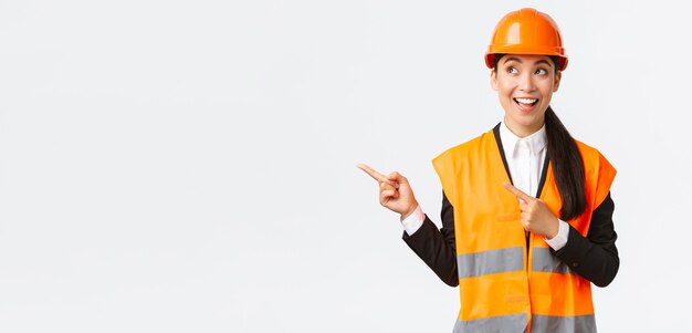 Building, construction and industrial concept. Excited and interested smiling asian architect, engineer in helmet and safety clothing looking, pointing upper left corner at interesting project