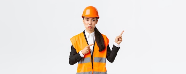 Building construction and industrial concept Upset and disappointed female asian manager engineer in safety helmet pouting complaining something bad pointing fingers upper right corner