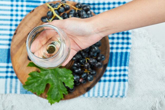 A bunch of black grapes on wooden plate with leaf while hand holding an empty glass