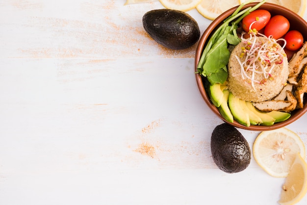 Burrito bowl with chicken; spinach; tomato; avocado and lemon slices in bowl on white wooden textured backdrop