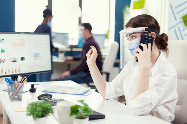 Free Photo business administrator sitting at her workplace wearing face mask during covid19 talking on smartphone. multiethnic coworkers working respecting social distance in financial company.