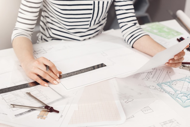 Business concept. Close up detail of young successful architect entrepreneur in striped clothes sitting at white table, looking through work plan, holding pen and ruler in hands, working in new busine