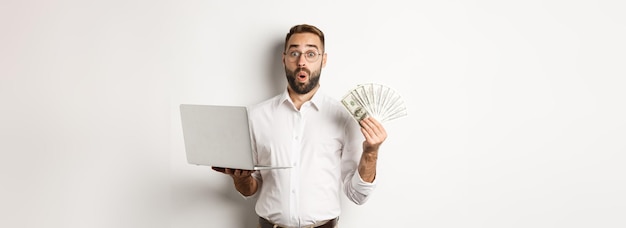 Free photo business and ecommerce man looking amazed with money income working online using laptop standing ove