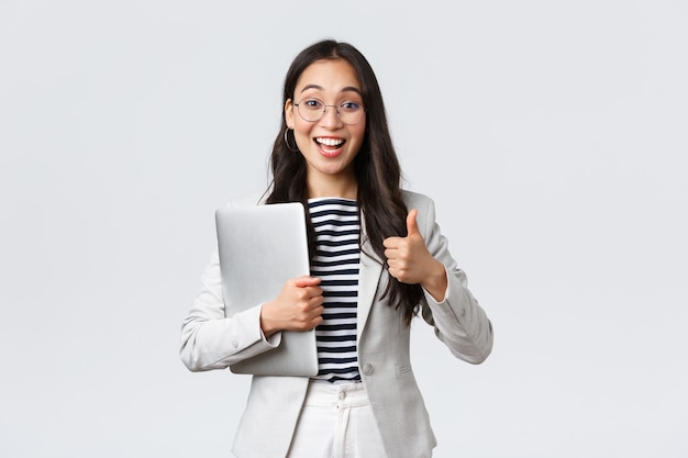 Business, finance and employment, female successful entrepreneurs concept. Young confident businesswoman in glasses, showing thumbs-up gesture, hold laptop, guarantee best service quality