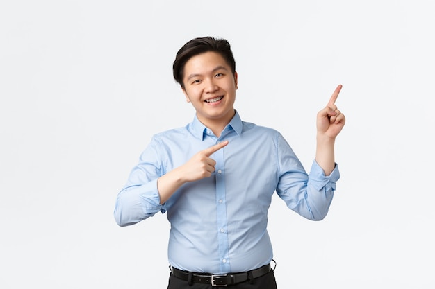 Business, finance and people concept. Pleasant smiling asian salesman in blue shirt, teeth braces, pointing fingers upper right corner, making announement, showing chart or product, white background.