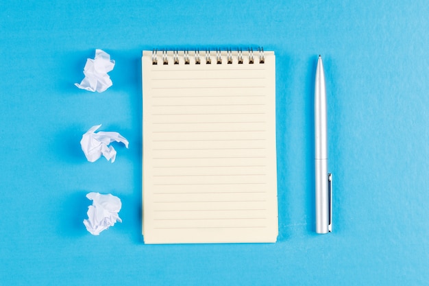 Business and financial concept with crumpled paper wads, spiral notebook, pen on blue background flat lay.