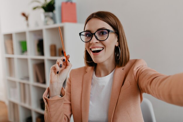 Business lady with glasses and in stylish light outfit makes selfie, holding orange pencil in her hands.