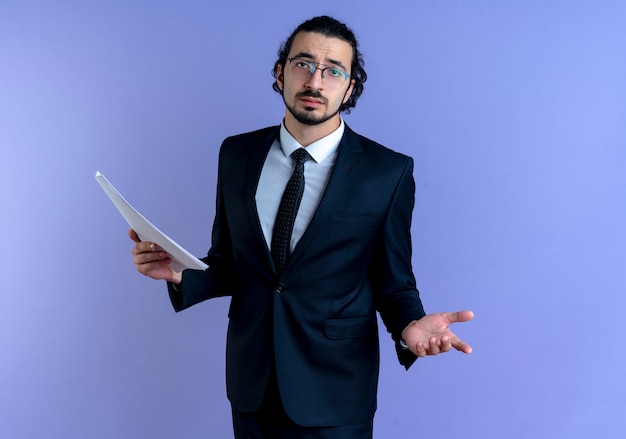 Business man in black suit and glasses holding documents looking to the front confused standing over blue wall