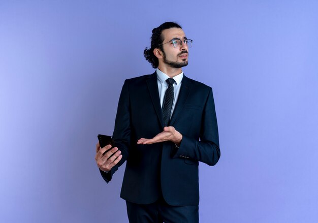 Business man in black suit and glasses holding smartphone looking confused standing over blue wall