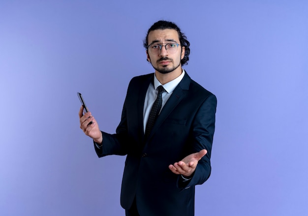 Business man in black suit and glasses holding smartphone looking to the front with arm out as asking standing over blue wall