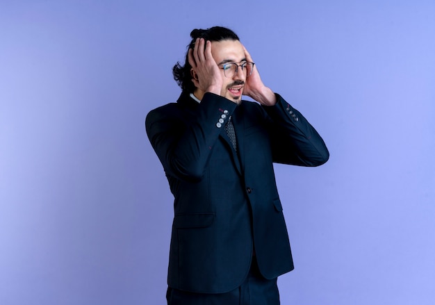 Business man in black suit and glasses looking aside holding his head with hands confused and very anxious standing over blue wall