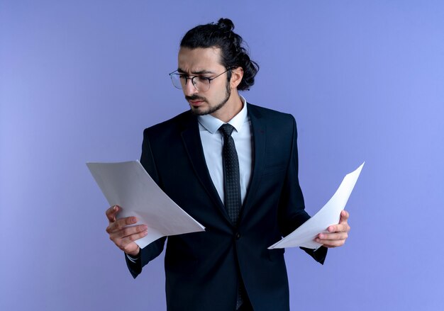 Business man in black suit and glasses looking at documents with serious face standing over blue wall