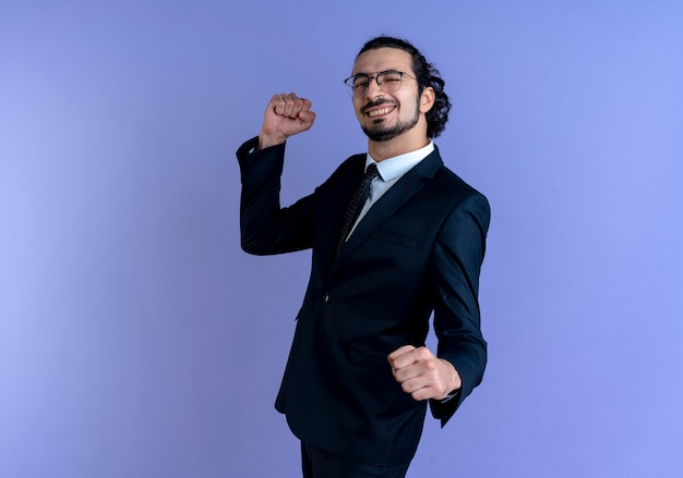 Business man in black suit and glasses looking to the front clenching fists happy and excited standing over blue wall