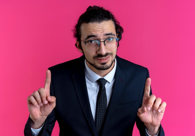 Free photo business man in black suit and glasses looking to the front showing index fingers standing over pink wall