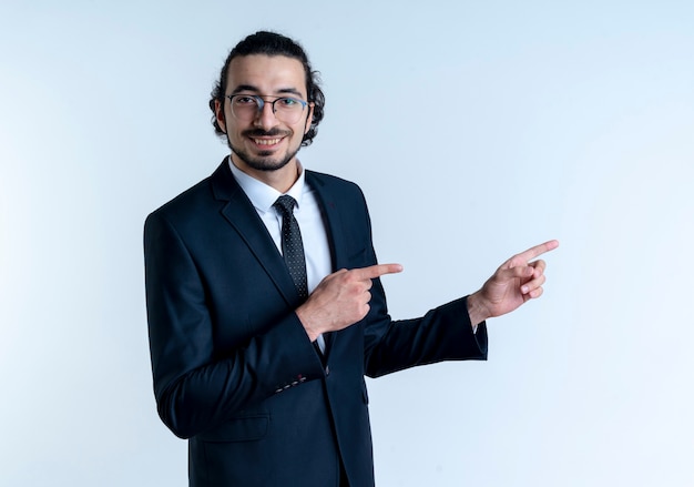 Business man in black suit and glasses looking to the front smiling cheerfully pointing with index fingers to the side standing over white wall