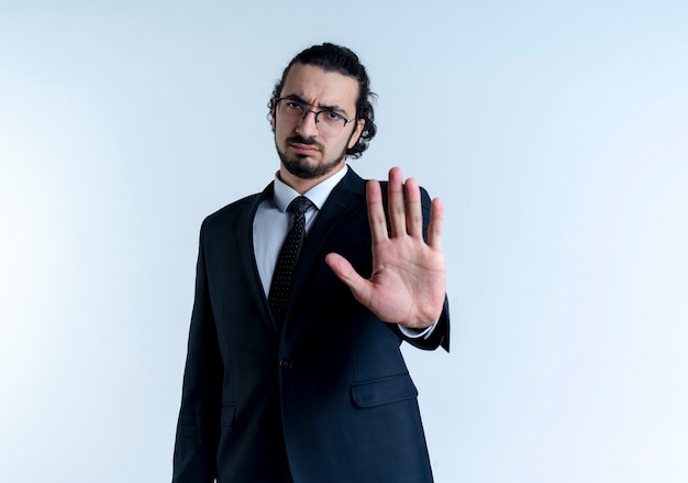 Business man in black suit and glasses making stop sign with open hand looking to the front with serious face standing over white wall