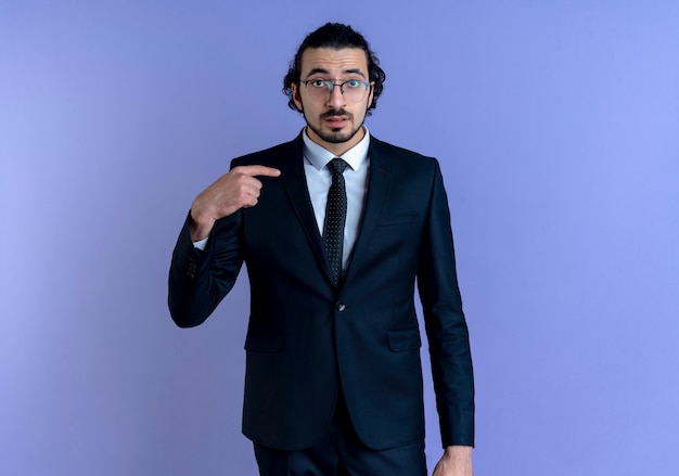 Free Photo business man in black suit and glasses pointing with finger to himself looking confused standing over blue wall