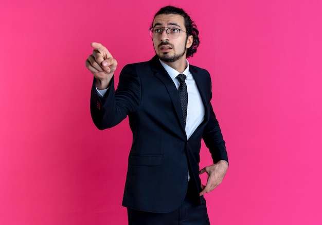 Free Photo business man in black suit and glasses pointing with finger to the side looking surprised standing over pink wall