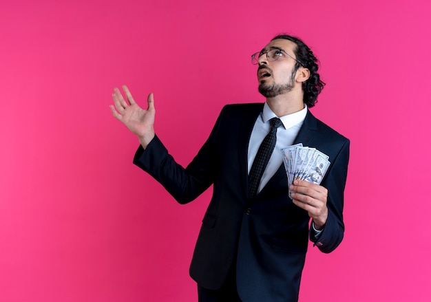 Free photo business man in black suit and glasses showing cash, looking up confused with arm out standing over pink wall