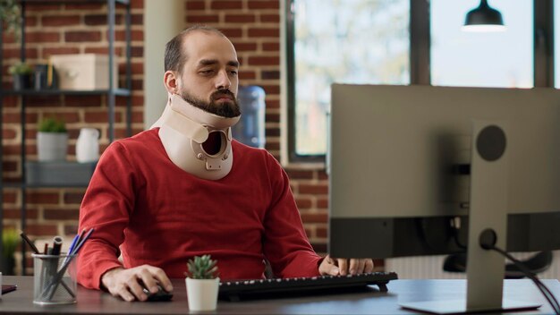 Business man using neck collar to recover from physical injury, working on financial strategy charts and graphs. Office employee wearing cervical foam to cure fracture at job.