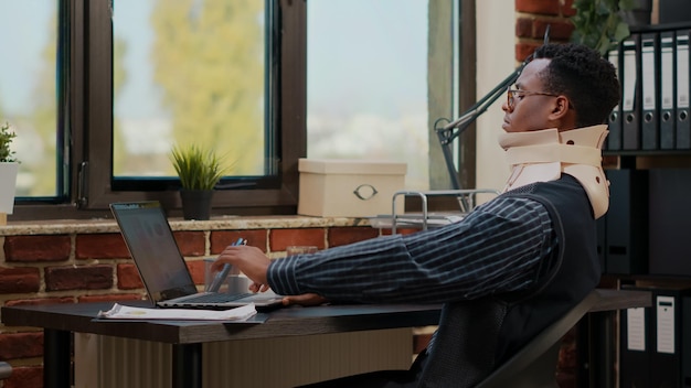 Business man wearing cervical neck collar at work after vertebrae injury in startup office. Injured employee using medical foam while he works with laptop to plan commercial investment.