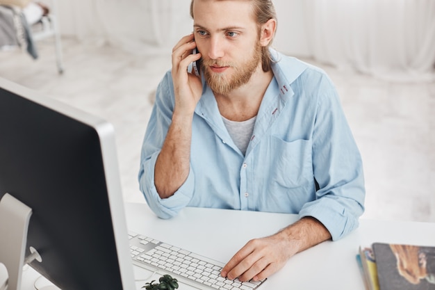 Business, office and technology concept. Top view of bearded employee wearing blue shirt, talking on phone with companions, typing on keyboard, looking on computer screen, using modern devices