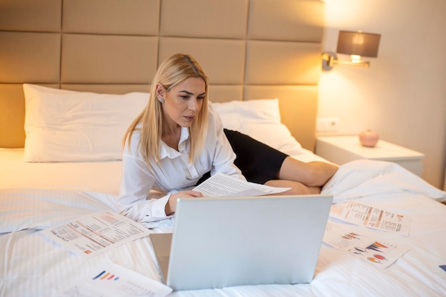 Free photo business technology internet and hotel concept happy businesswoman with laptop computer lying in hotel in bed and working late in hotel room with her laptop computer document and paperwork